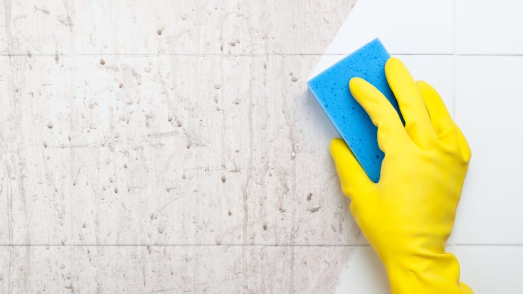 gloved hand holding a sponge and wiping away dirt and grime from a tiled wall