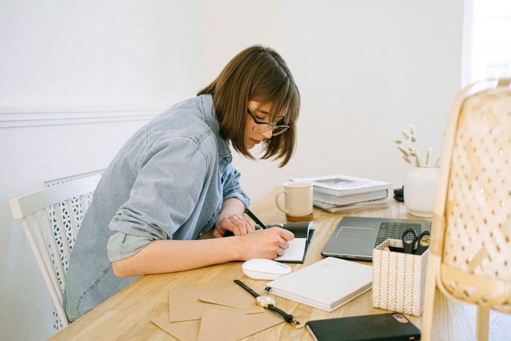 woman sitting at desk in front of laptop writing in notebook
