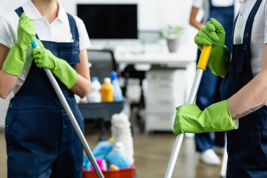 Two cleaners in a white pollo shirt and navy blue overalls wearing gloves as they mop the floor