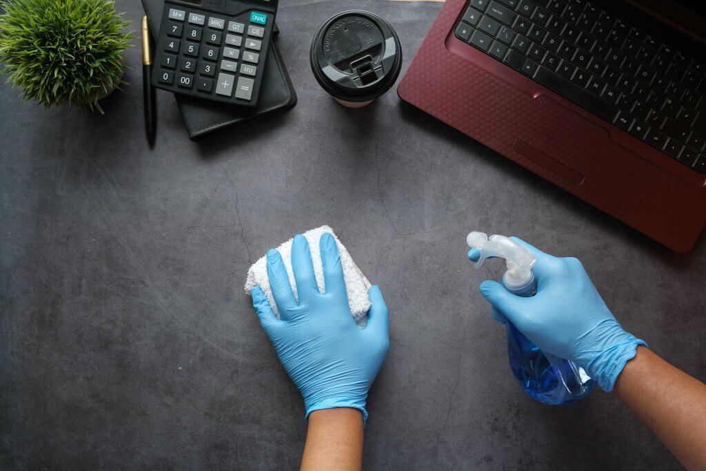 gloved hands with a spray bottle and towel cleaning desk with laptop and calculator off to the side