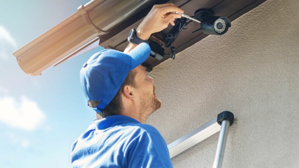 Man in a blue baseball cap and blue polo is installing a surveillance camera on the outside of a home