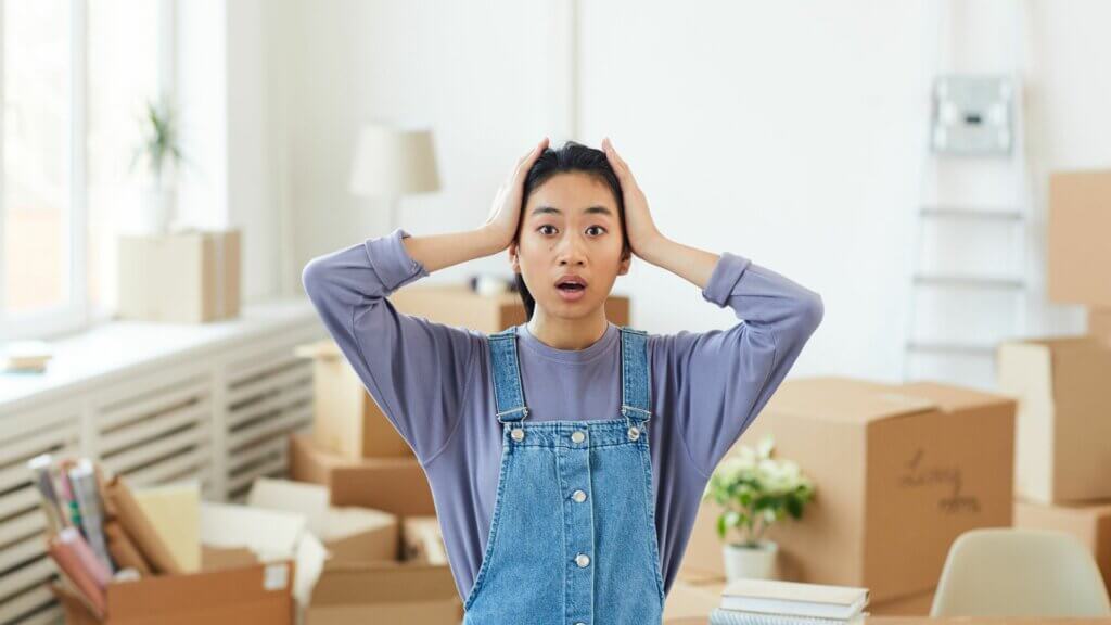 Airbnb cleaner with a shocked expression holding hands on either side of her head with brown boxes behind her. 