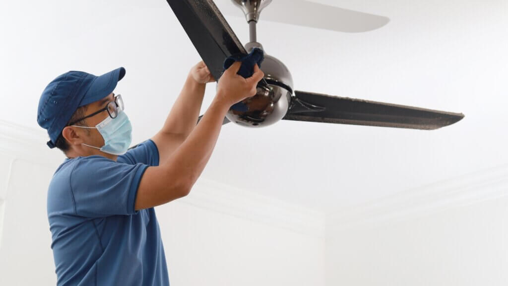 Male cleaning crew member wearing a blue medical mask cleaning dust off ceiling fan with microfiber cloth