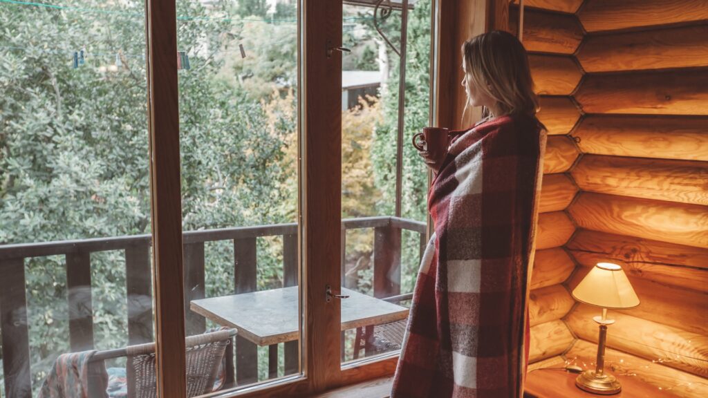 woman holding a mug wrapped in a plaid blanket looking out cabin bedroom window 